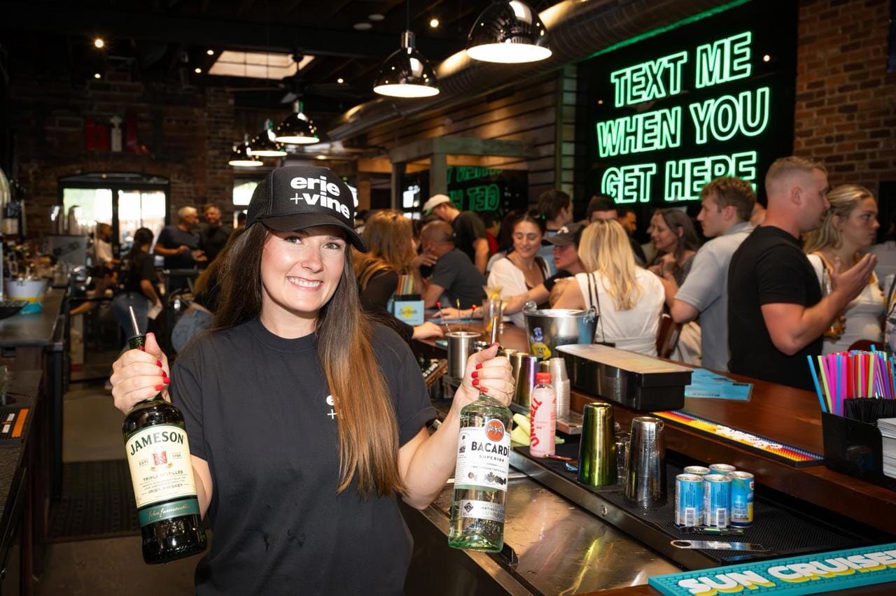 Smiling bartender holding bottles of Jameson and Bacardi behind a packed bar at Erie + Vine in downtown Willoughby, Ohio, with a neon sign that reads “Text Me When You Get Here.”