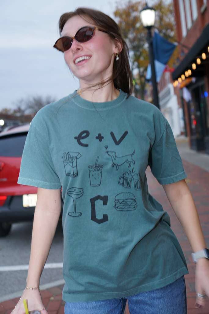 Woman wearing Erie + Vine's signature green tee with illustrations of fries, iced coffee, dog-friendly vibes, cocktails, and burgers—representing the Willoughby bar's unique identity.